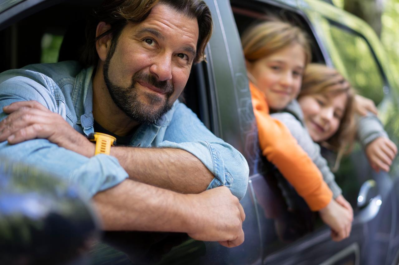 Homem sorridente no banco de motorista com os braços apoiados na janela do veículo estacionado, enquanto suas duas filhas pequenas sorridentes estão no banco de passageiro