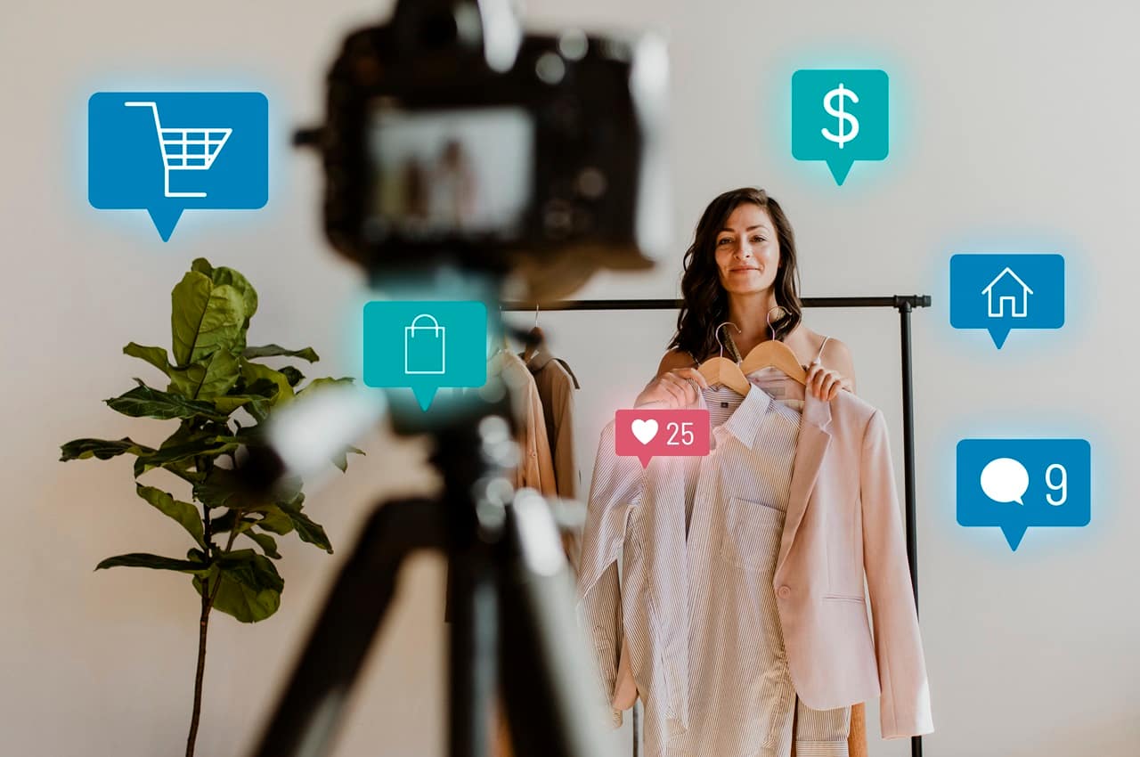 Mulher sorridente de cabelos castanhos longos vendendo camisa social e blazer rosa-claro em uma live ecommerce. Ela usa uma câmera no tripé para transmitir a live.