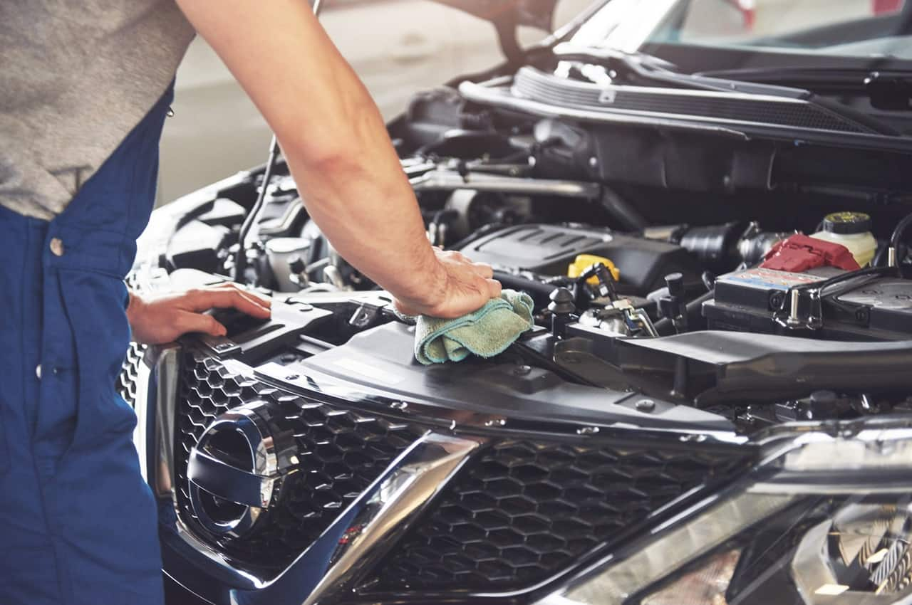 Mecânico veste macacão azul de segurança e faz vistoria veicular de um carro. O rosto da pessoa não aparece na foto e ele usa flanela verde para retirar tampa do recipiente de gasolina de um veículo. 