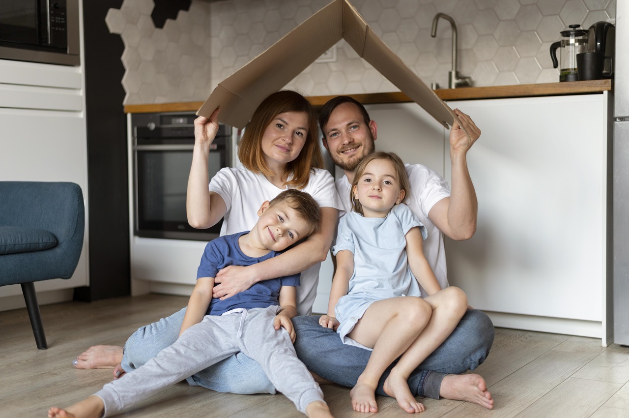 Família sorridente composta por um homem, uma mulher e duas crianças sentadas no chão. O casal segura um papelão em cima da cabeça de todos simulando o telhado de uma casa.