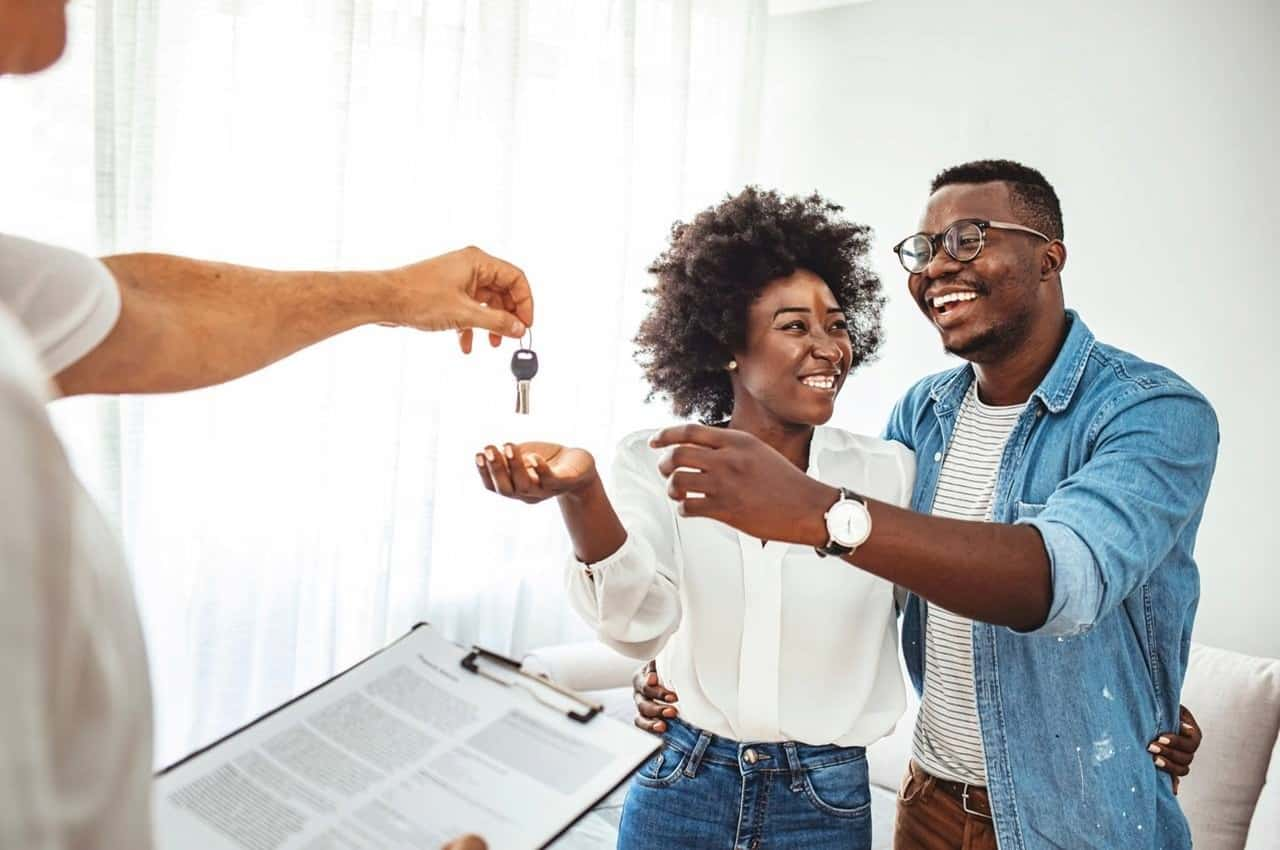 Casal sorridente recebendo chaves do novo apartamento com seguro. O vendedor segura nas mãos o contrato do apartamento e entrega as chaves para a mulher e o homem a sua frente.