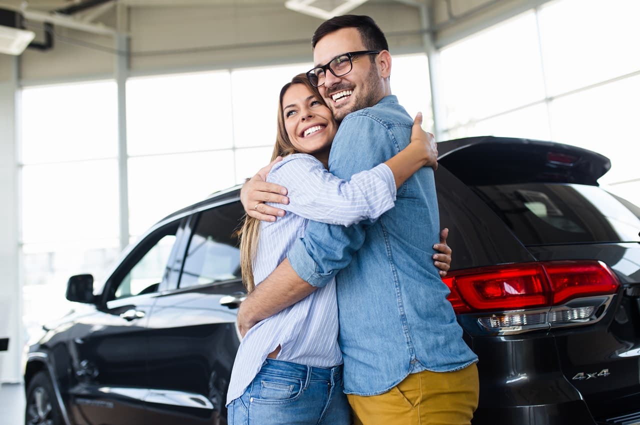 Casal sorridente abraçado em frente a um veículo SUV de cor preta. A mulher veste calça jeans e blusa social listrada azul. O homem veste blusa jeans e calça cáqui.