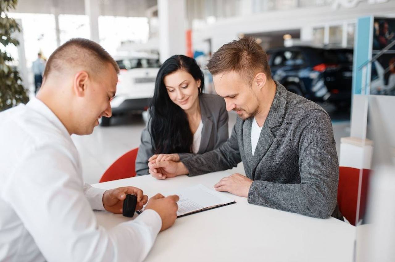 Casal sorridente (homem e mulher) assinando um contrato de veículo junto ao vendedor. Eles estão sentados em uma mesa dentro da concessionária de carros.