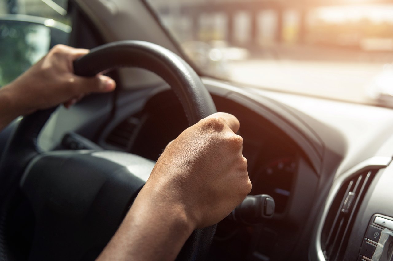 Mãos segurando volante básico de carro, enquanto dirige. A pessoa não aparece na imagem.