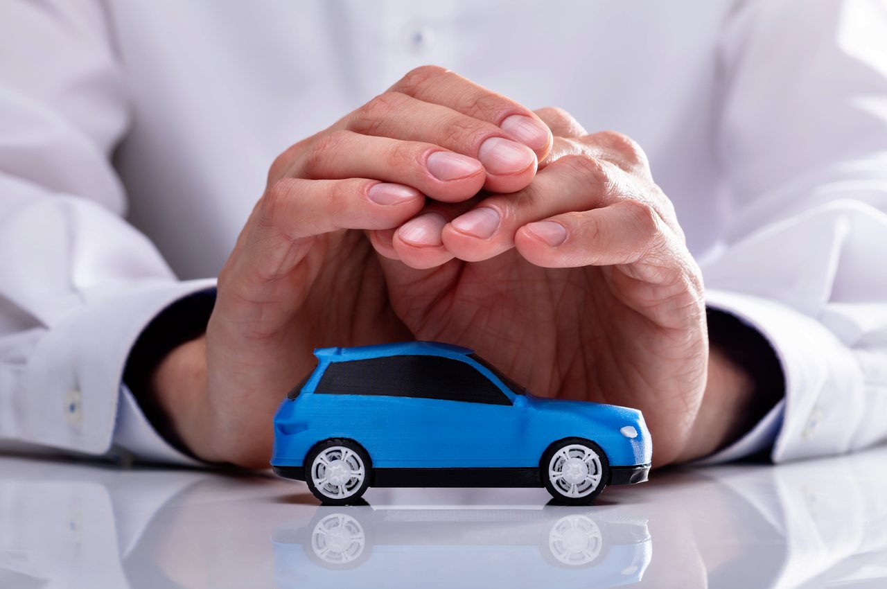 Pessoa (seu rosto não aparece na imagem) utilizando as mãos para proteger miniatura de carro azul em cima de uma mesa branca.