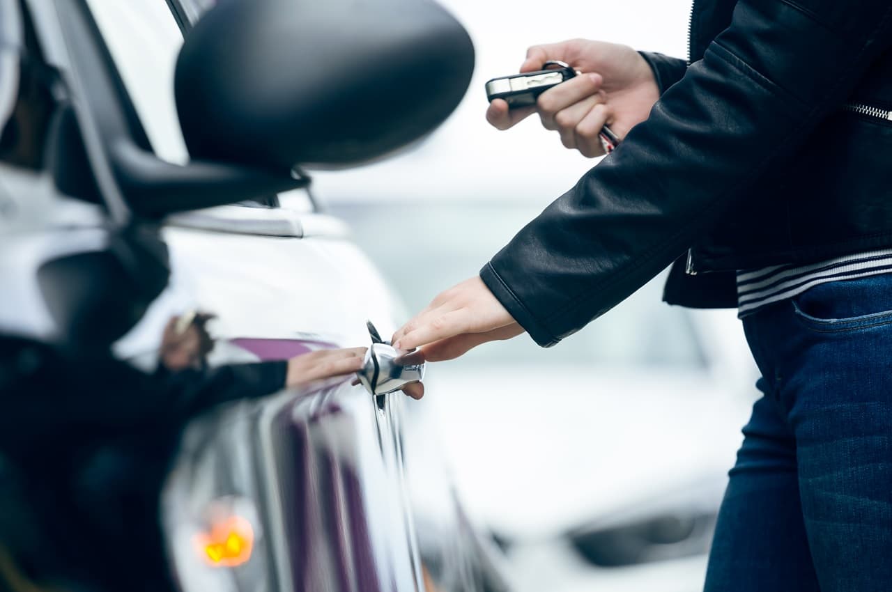 Pessoa em pé segurando maçaneta de carro e apontando o alarme para o veículo. A pessoa veste calça jeans, blusa listrada e casaco preto.