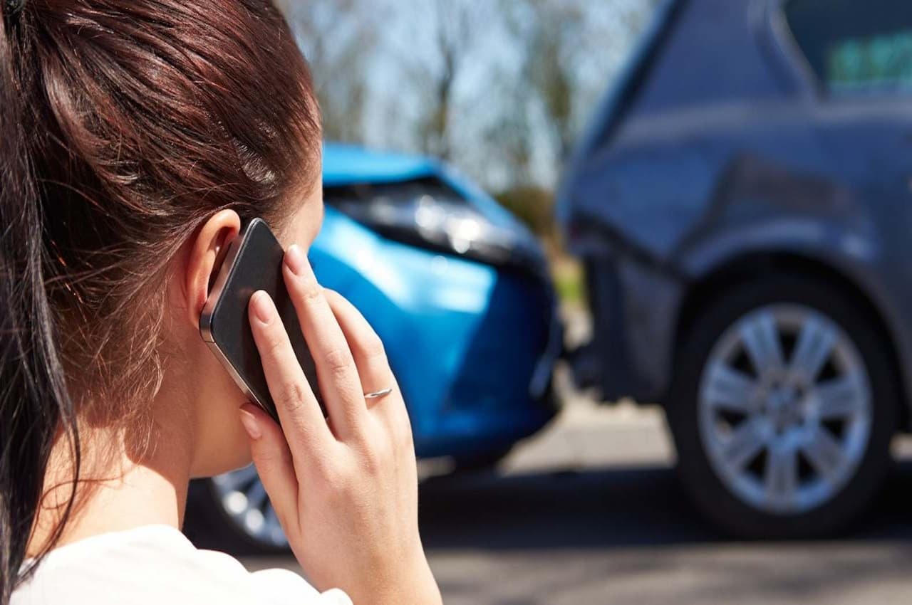 Mulher de cabelos presos castanhos, ligando no celular para seguro e proteção veicular após acidente de carro. Ela observa seu veículo batido na traseira de outro.