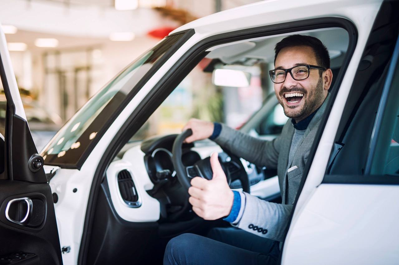 Homem sorridente, sentado no banco de motorista de um veículo que está na concessionária. Ele faz um OK com as mãos e está com a porta do veículos aberta.