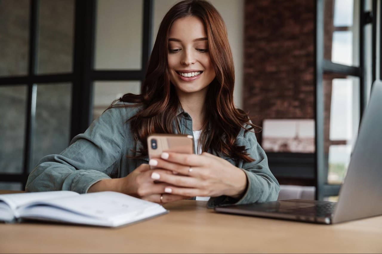 Mulher sorridente com cabelos longos castanhos, sentada no escritório e utilizando celular, caderno e computador para calcular valor do seguro de carro.