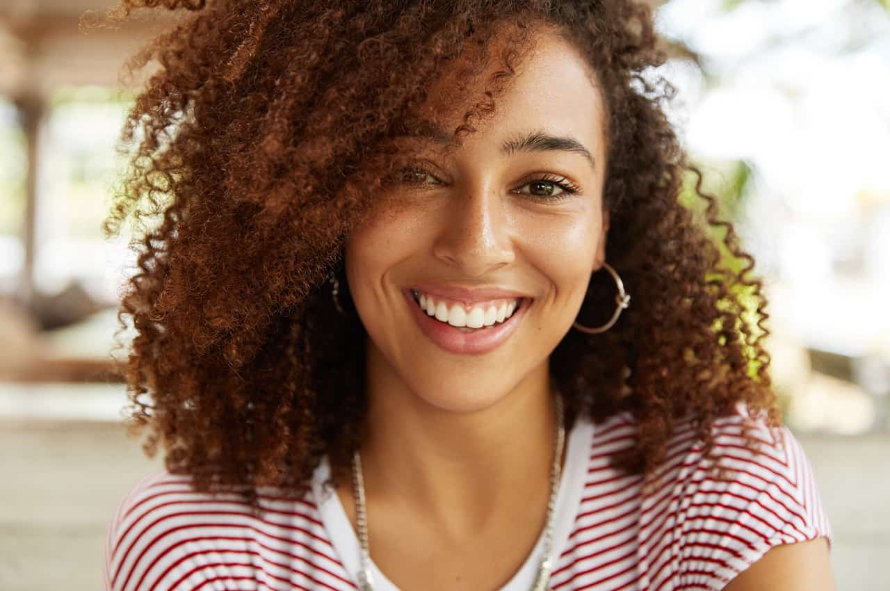 Close-up em rosto de mulher negra sorridente com cabelos soltos e brincos de argola. Ela veste camisa listrada vermelha e branca e um colar prata