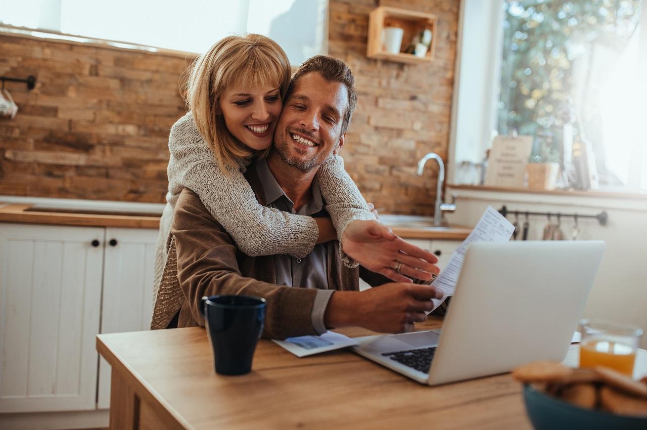 Mulher abraçando homem sorridente que está segurando folha de contas do mês e utilizando um computador na cozinha da sua casa.