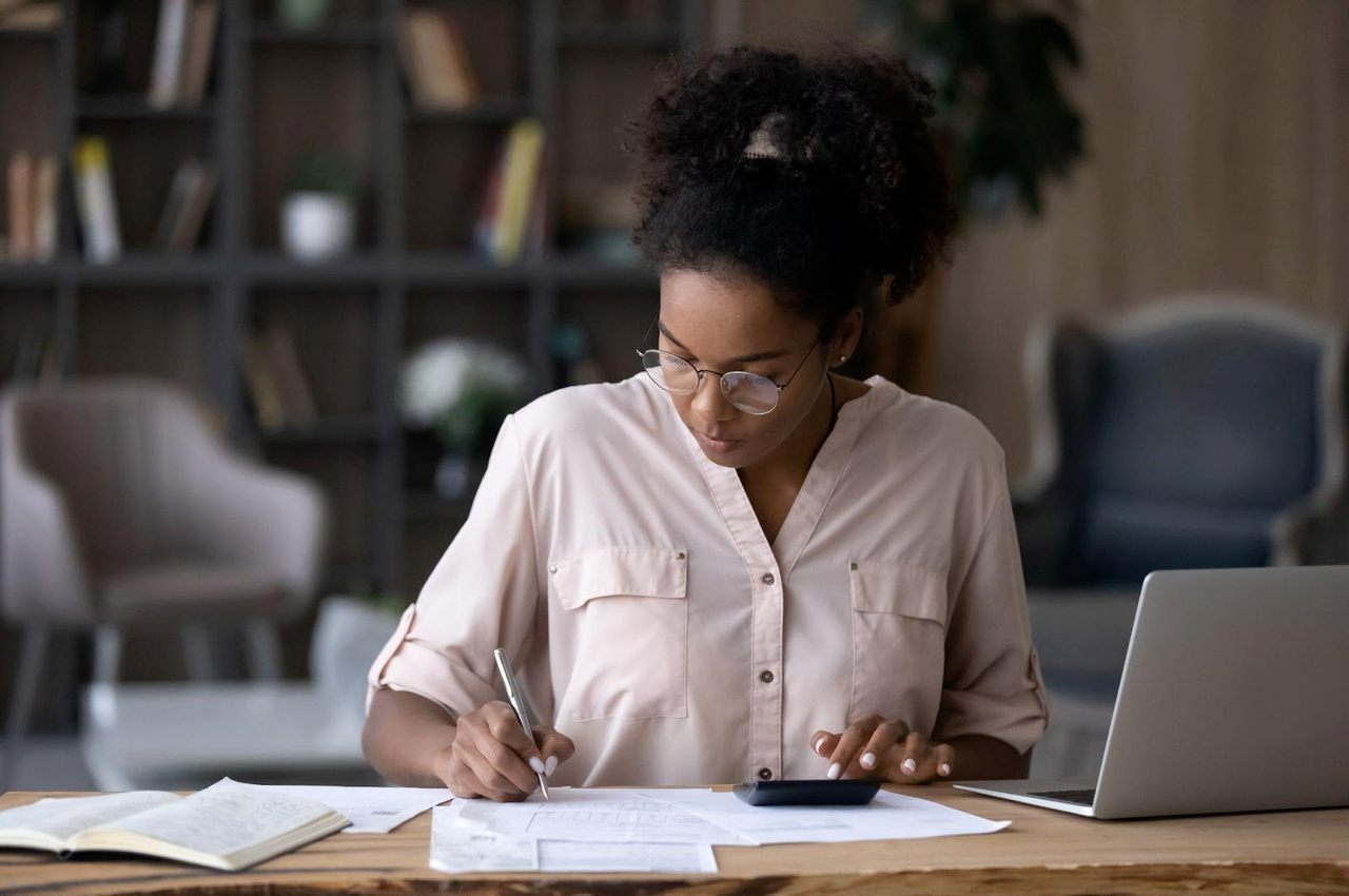 Mulher com expressão concentrada realizando cálculo de custo de vida com uma calculadora e anotando em papéis sob a mesa do escritório.