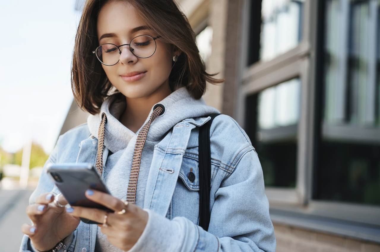 Mulher sorridente de cabelos castanhos curtos, óculos de grau e jaqueta jeans com moletom, acessando banco digital pelo celular na rua.