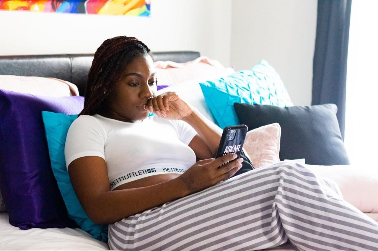 Mulher com expressão concentrada acessando canais digitais bancários pelo celular. Ela está sentada na cama, apoiada em almofadas azuis e roxas, vestindo um pijama listrado.