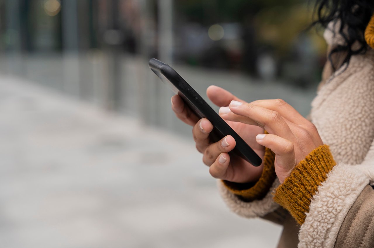 Foco em pessoa utilizando celular para acessar boleto online. O rosto da pessoa não aparece na imagem e ela veste casaco de pelos.