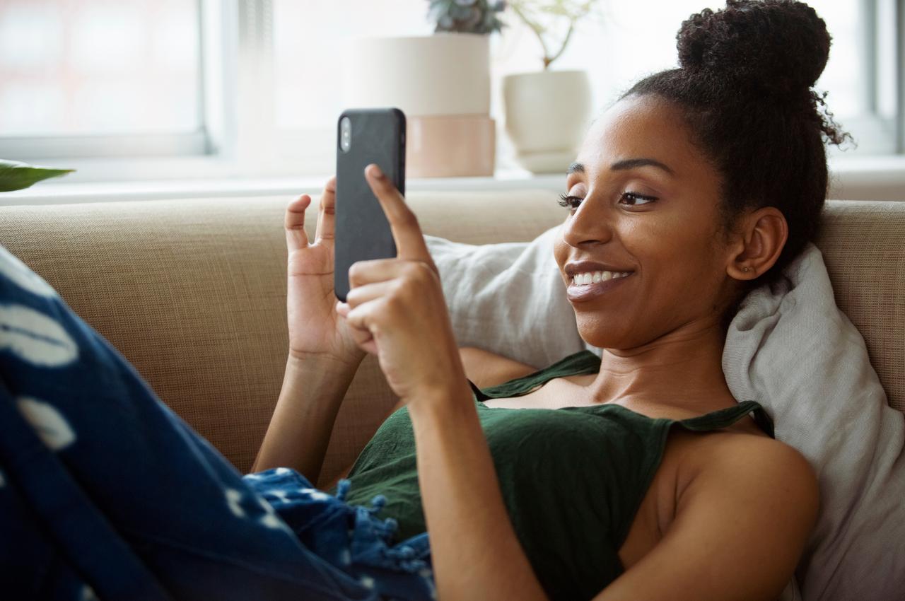 Mulher sorridente deitada no sofá da sala utilizando celular para acessar conta-corrente. Ela veste regata verde, calça jeans e coque no cabelo.