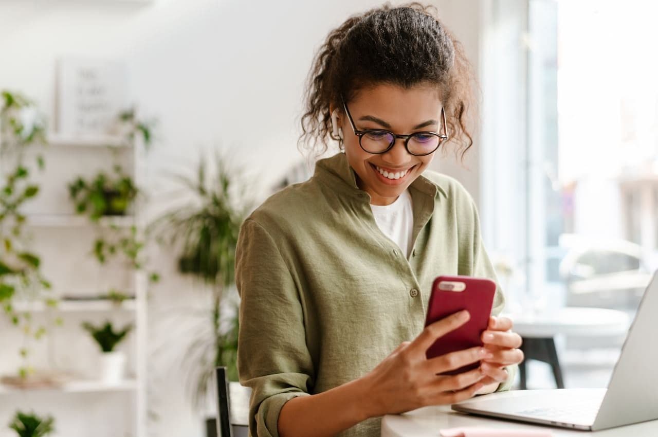 Mulher sorridente com óculos de grau, sentada em frente ao seu computador e acessando débito automático pelo celular. Ela veste camisa de botões verde-musgo.