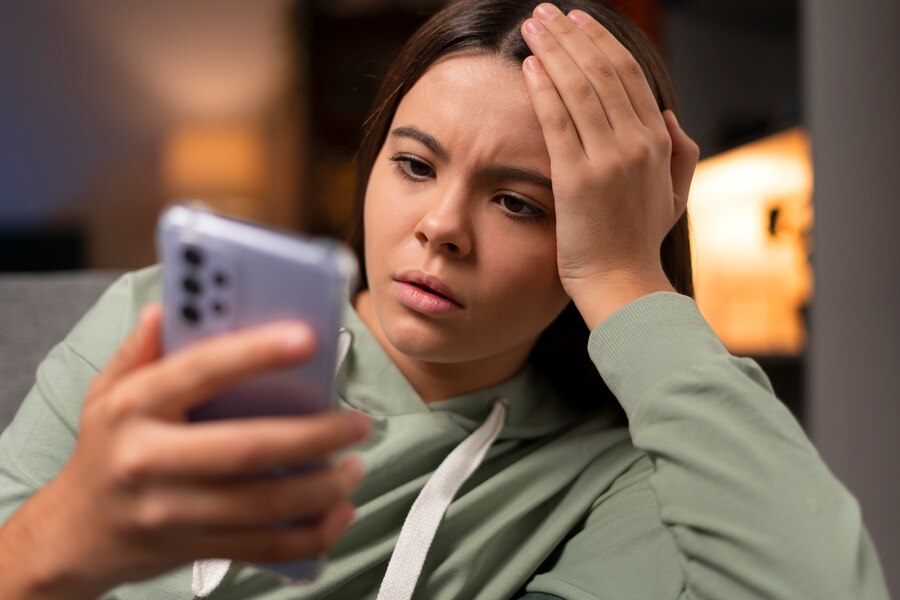 Mulher jovem segurando um celular com uma das mãos e a outra leva à cabeça, em gesto de preocupação. Ela usa um moletom verde musgo.