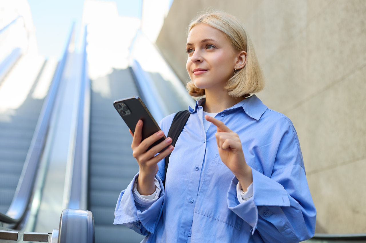 Mulher loira sorridente, segurando um smartphone. Ela está em frente a uma escada rolante, vestindo uma camisa azul, em um ambiente urbano ao ar livre.