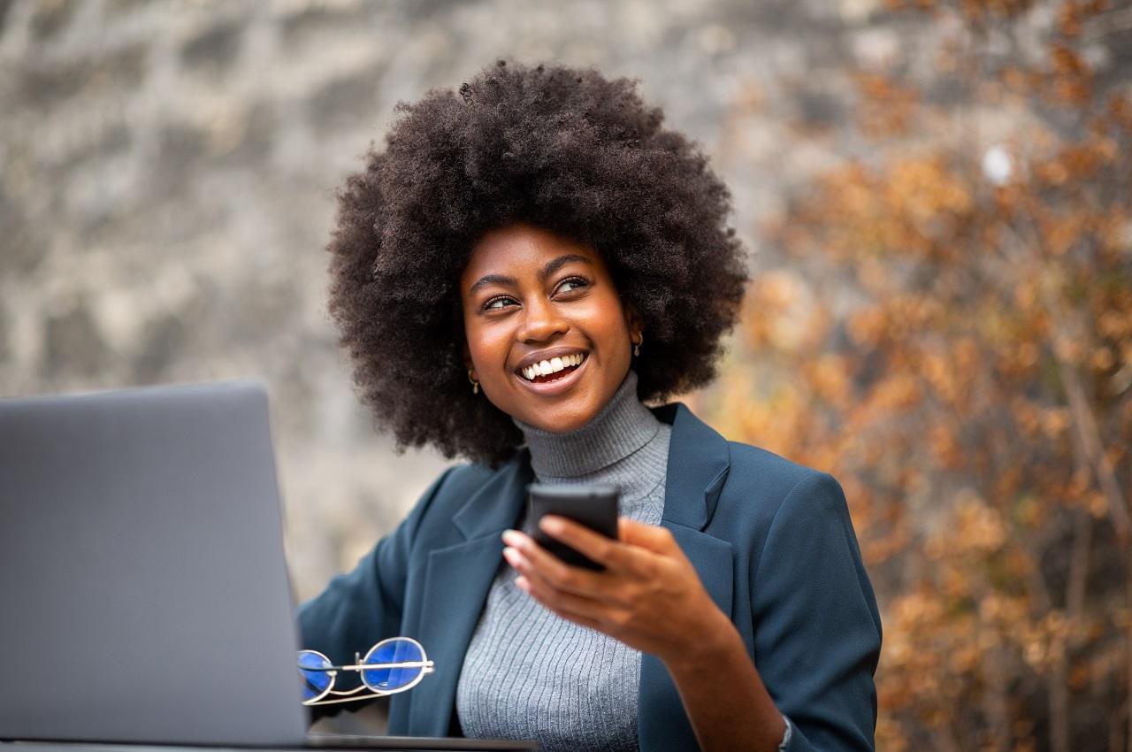 Mulher sorridente sentada em uma praça arborizada e utilizando celular para acessar Pix. Em cima da mesa da praça, tem um laptop e um óculos de grau. A mulher veste blusa de lã e terno cinza.