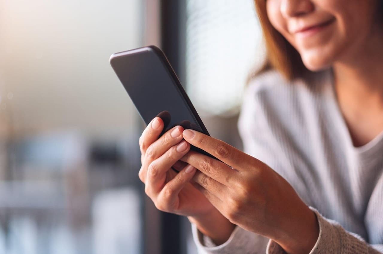 Foco em mulher segurando celular com ambas as mãos. Seu rosto e corpo aparecem borrados na imagem.