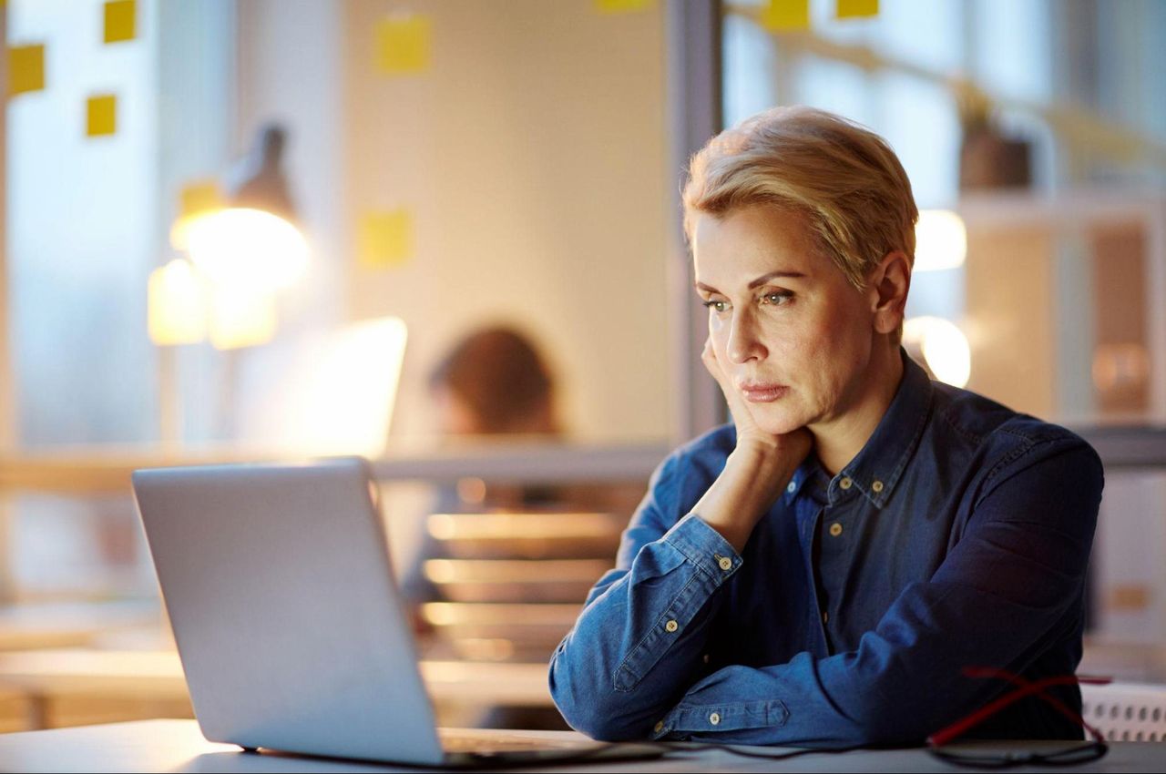 Mulher sênior com expressão concentrada, apoiando a mão esquerda no queixo e observando a tela de um notebook sob a mesa do escritório.