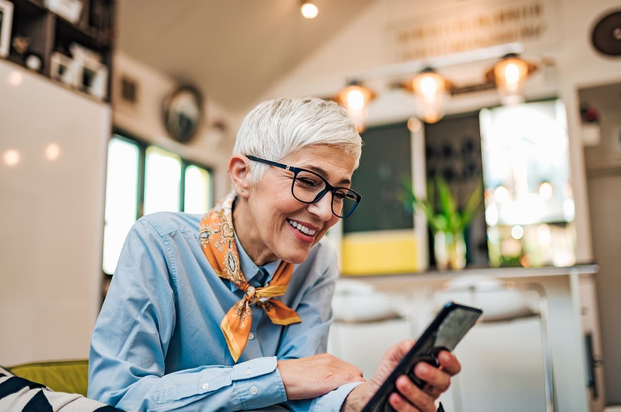 Mulher sênior sorridente com cabelos grisalhos. Ela veste um lenço amarelo-dourado no pescoço, blusa social azul-clara e acessa a conta digital pelo celular.