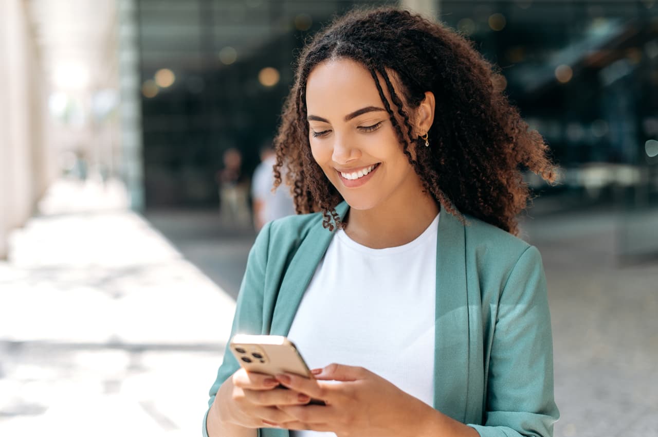 Mulher sorridente de cabelos cacheados soltos, veste blazer verde-água e digita como guardar dinheiro no celular.