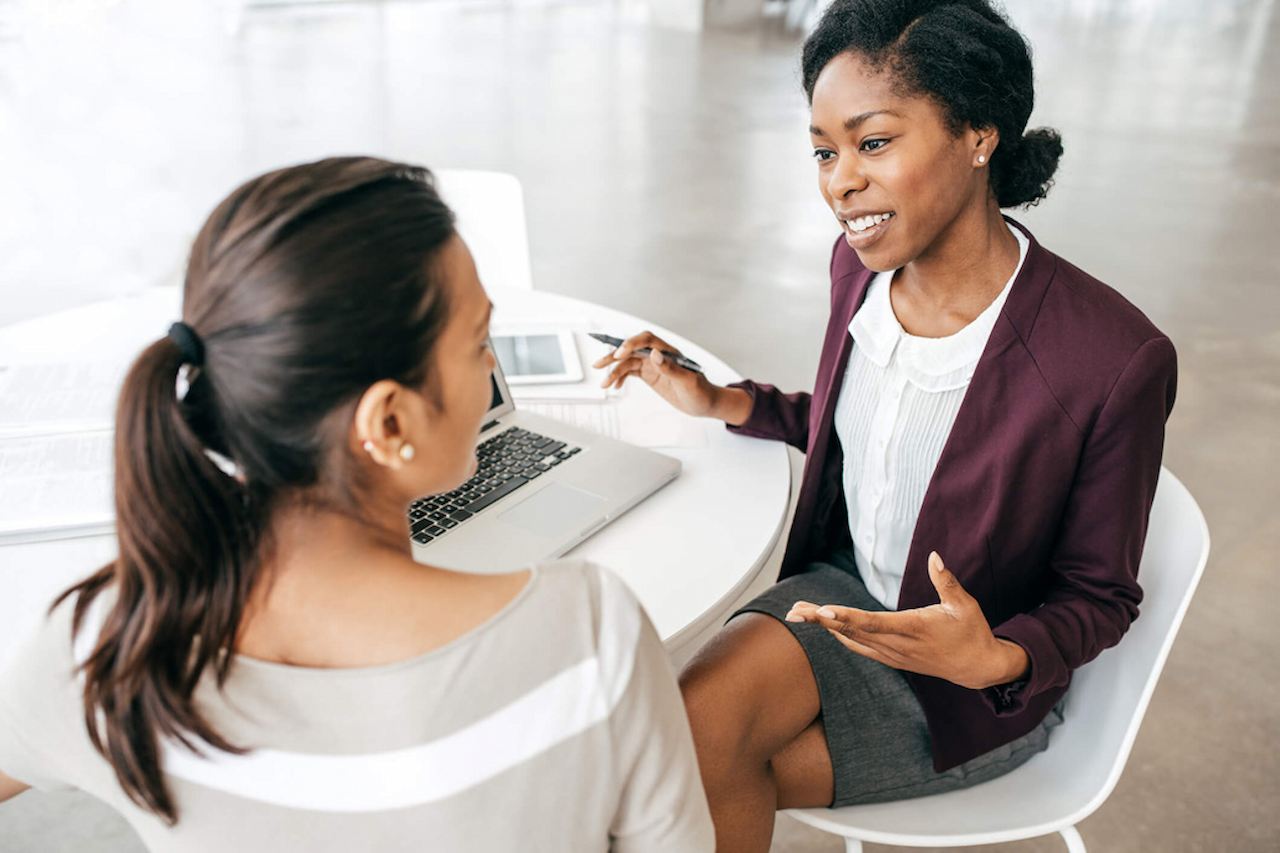 A imagem mostra duas mulheres sentadas à uma mesa, conversando. Uma delas está com um laptop à sua frente, segurando uma caneta.