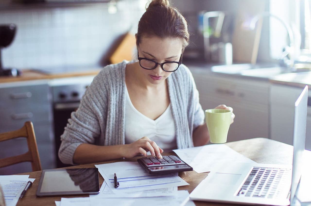 Mulher com expressão concentrada, segurando caneca de café e realizando contas de amortização de financiamento na calculadora. Em cima da sua mesa, tem um computador, tablet e anotações.