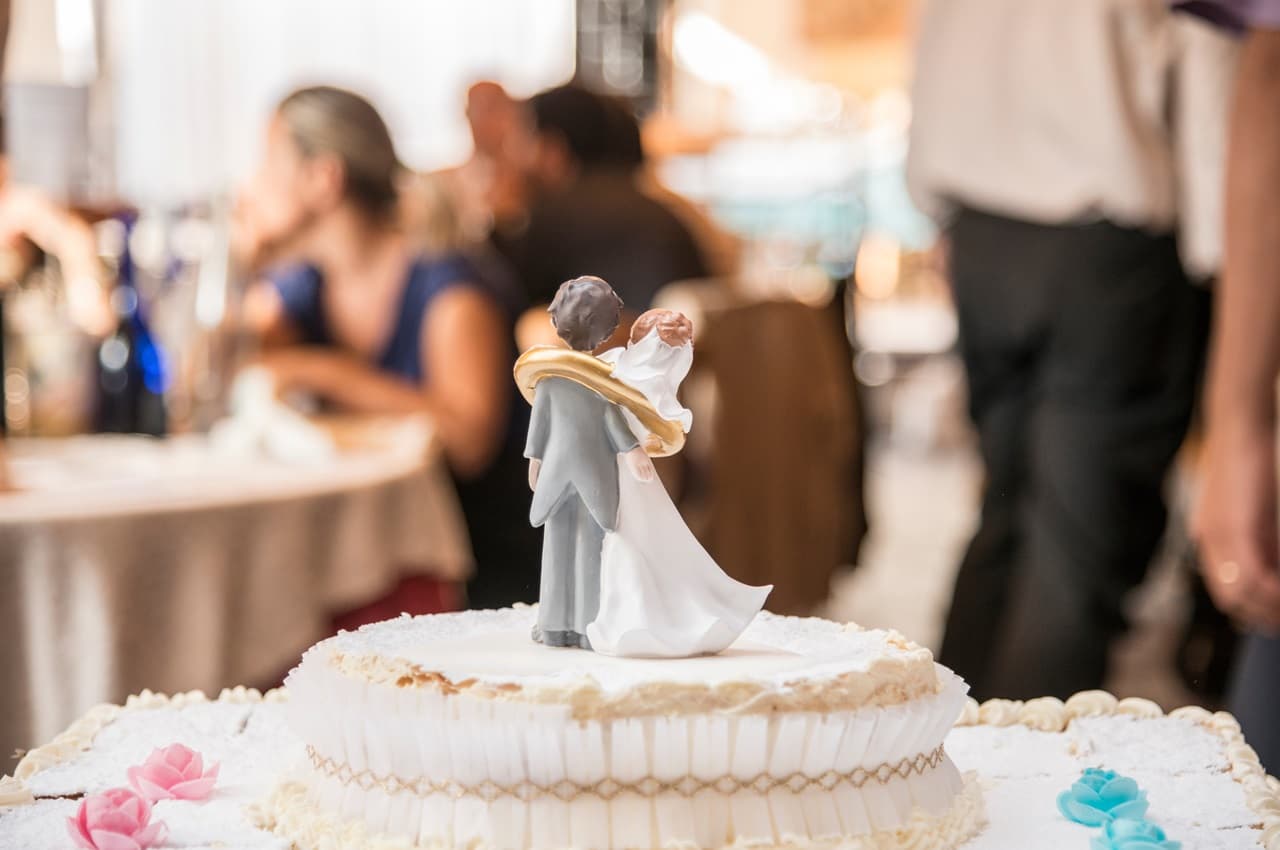 Topo de bolo com enfeite de noivinhos e, ao fundo, um salão de festa de aniversário de casamento desfocado.