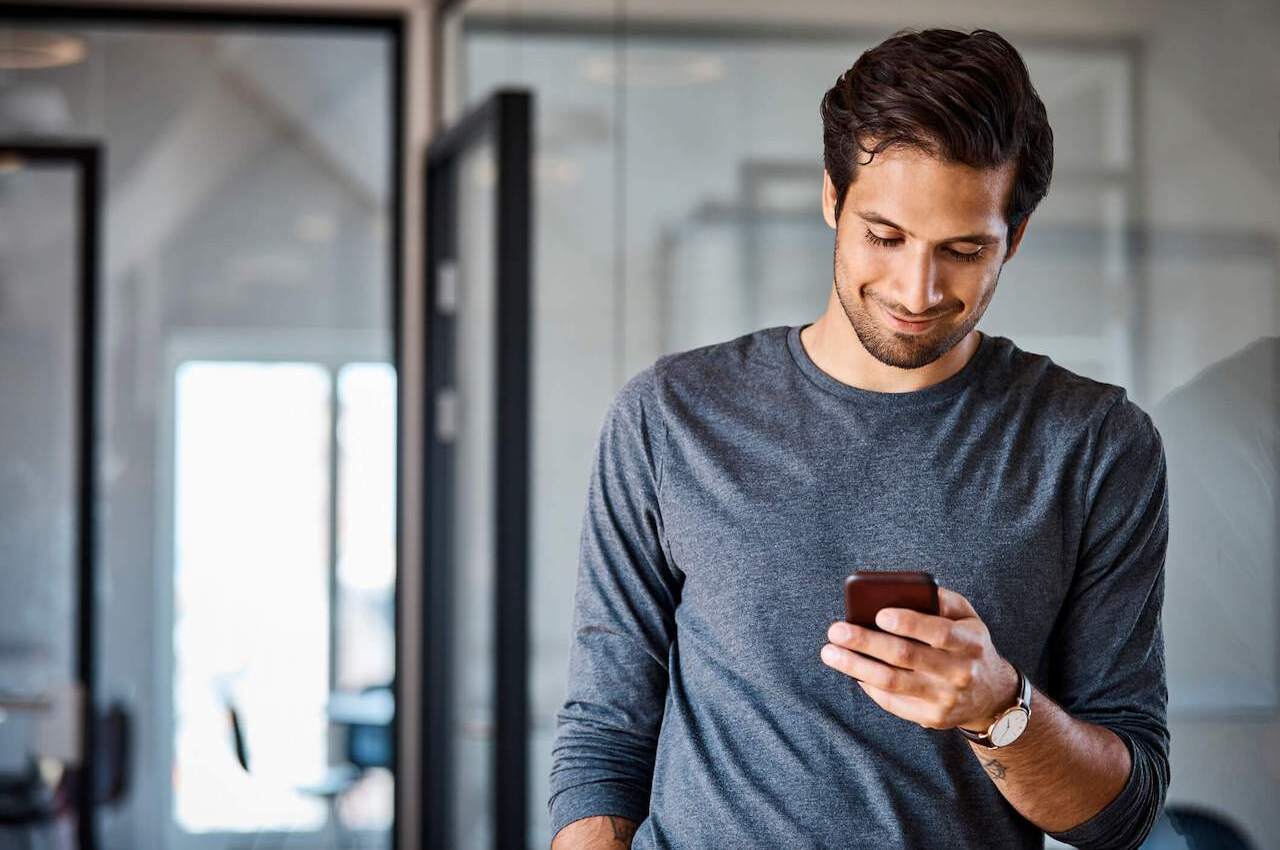 Homem sorridente, vestindo camisa cinza de mangas compridas dentro de casa e usando aplicativos para autônomos no celular.