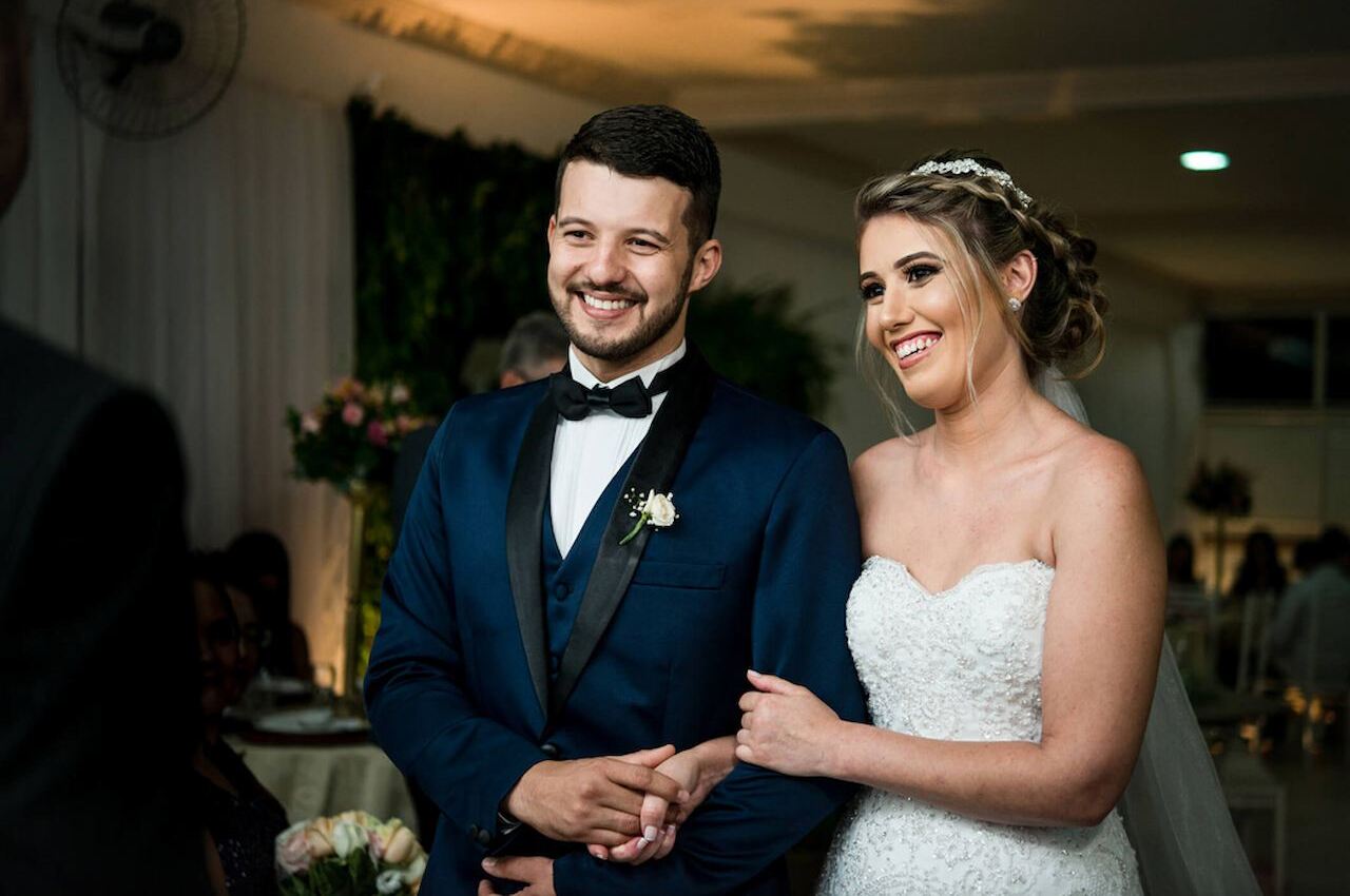 Casal de noivos sorridentes, composto por um homem de terno e uma mulher de vestido de noiva branco, no altar de casamento.