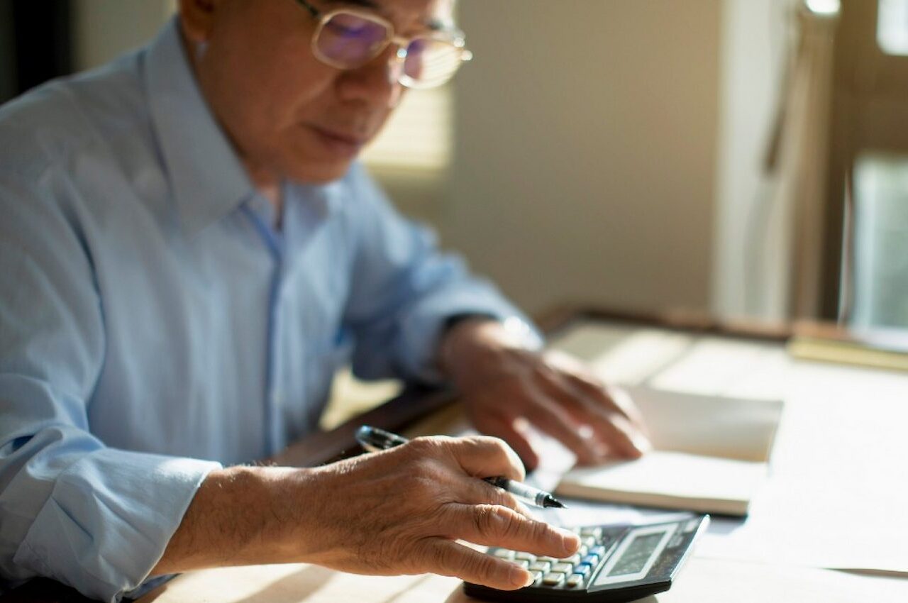 Homem sênior de cabelos grisalhos curtos e óculos de grau, utilizando calculadora para fazer cálculo da aposentadoria e anotando em um caderno.