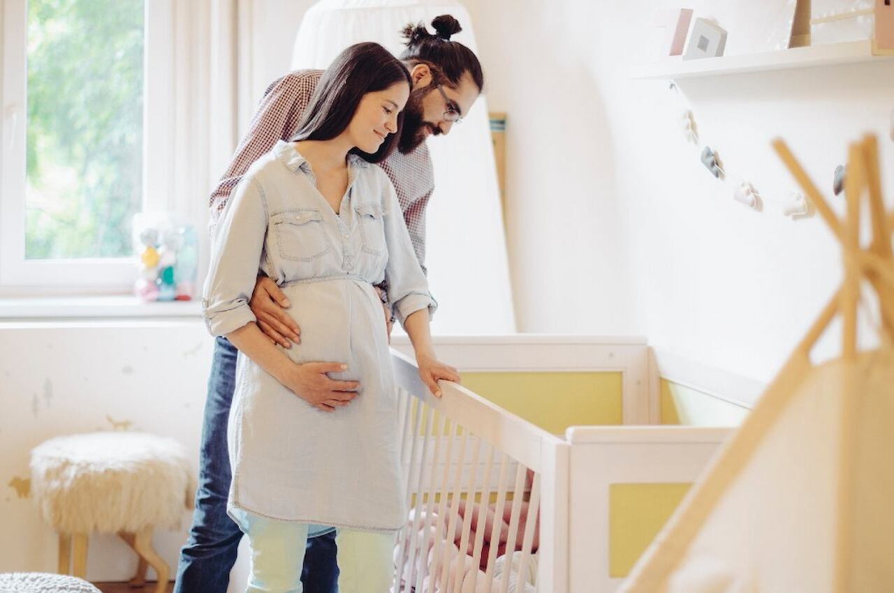 Casal sorridente com uma mulher grávida e seu companheiro, encostados no berço do futuro bebê. O quarto é iluminado, tem as paredes brancas e o berço um tom de amarelo claro.