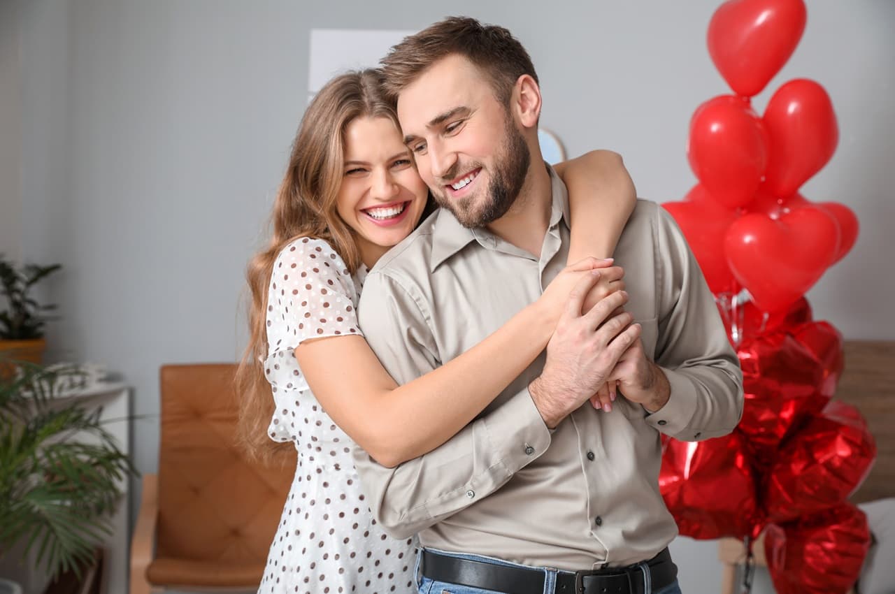 Casal sorridente composto por uma mulher loira abraçando seu companheiro de cabelos castanhos curtos no Dia dos Namorados. Ao fundo, tem balões vermelhos em formato de coração.