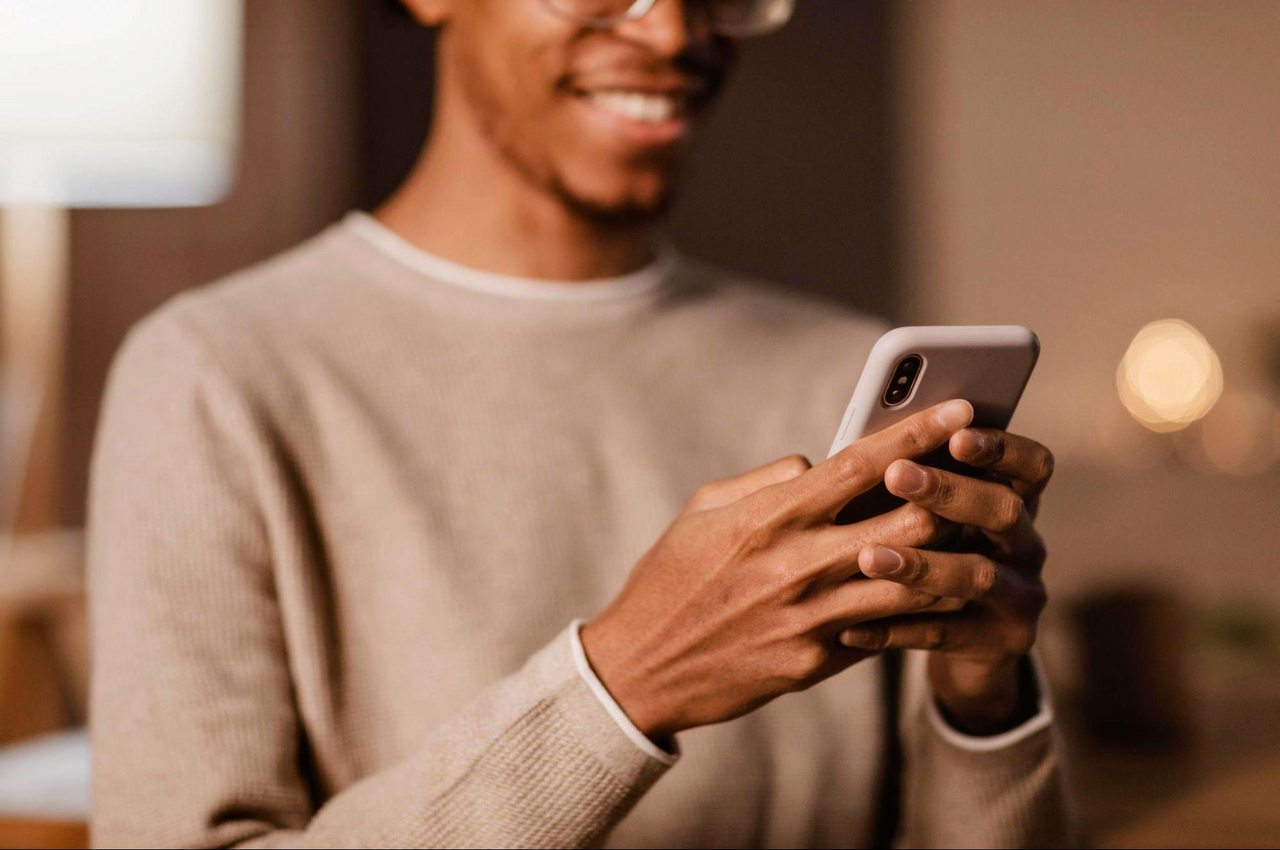 Homem sorridente, vestindo suéter marrom e digitando no celular como declarar imposto de renda.