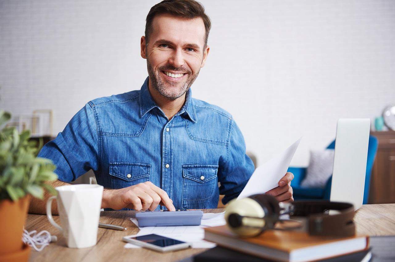 Homem sorridente de cabelos curtos castanhos e blusa jeans, utilizando uma calculadora e segurando uma folha branca A4.