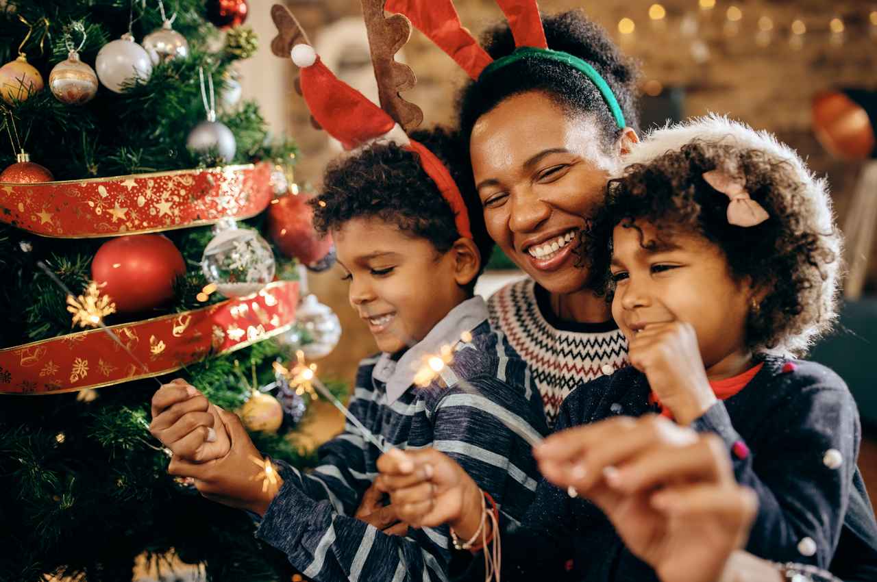 Família sorridente (mulher abraçada com duas crianças pequenas) segurando velas comemorativas próxima a árvore de natal