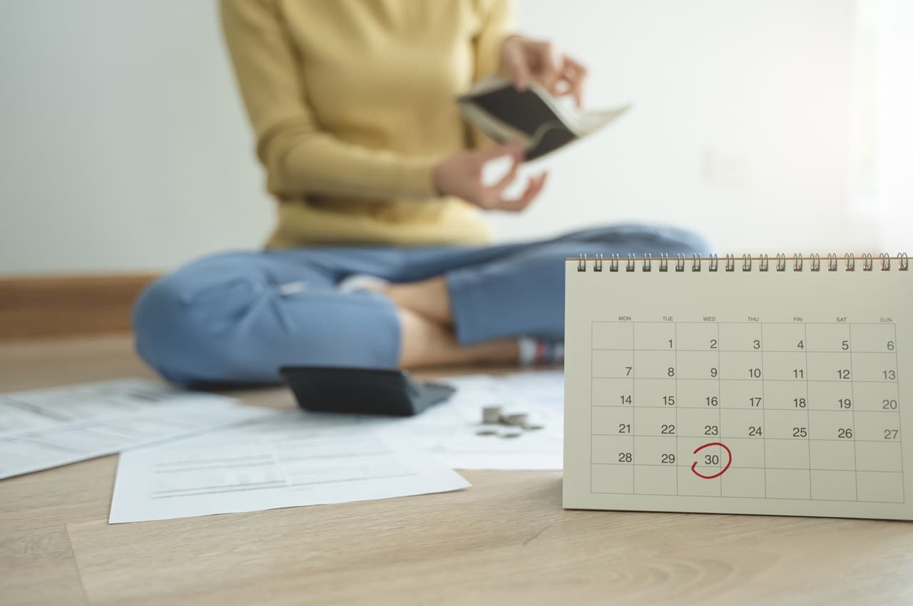 Pessoa (o rosto não aparece na imagem) sentada no chão com as pernas cruzadas e uma carteira vazia aberta, enquanto tem contas atrasadas espalhadas pelo chão e um calendário riscado.