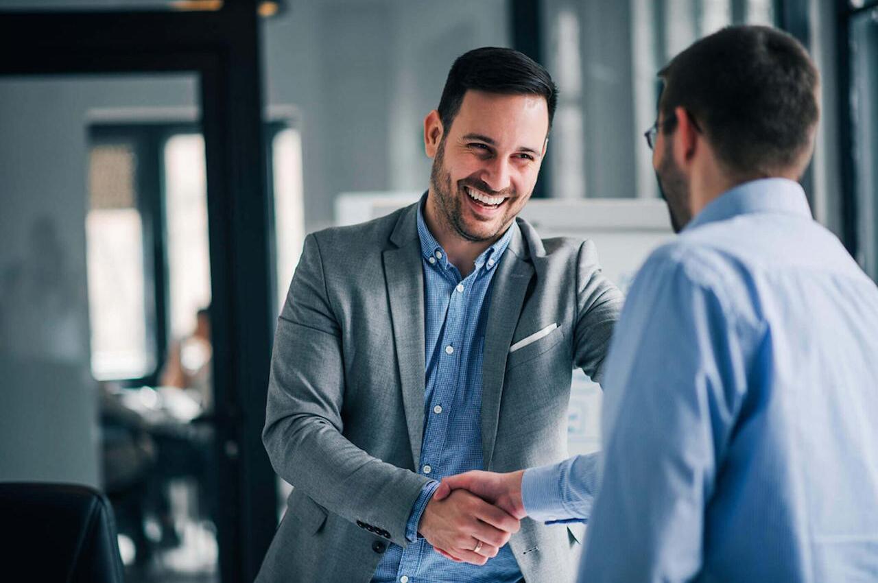 Dois homens sorridentes, vestindo ternos cinzas e apertando as mãos em cumprimento no ambiente de trabalho.