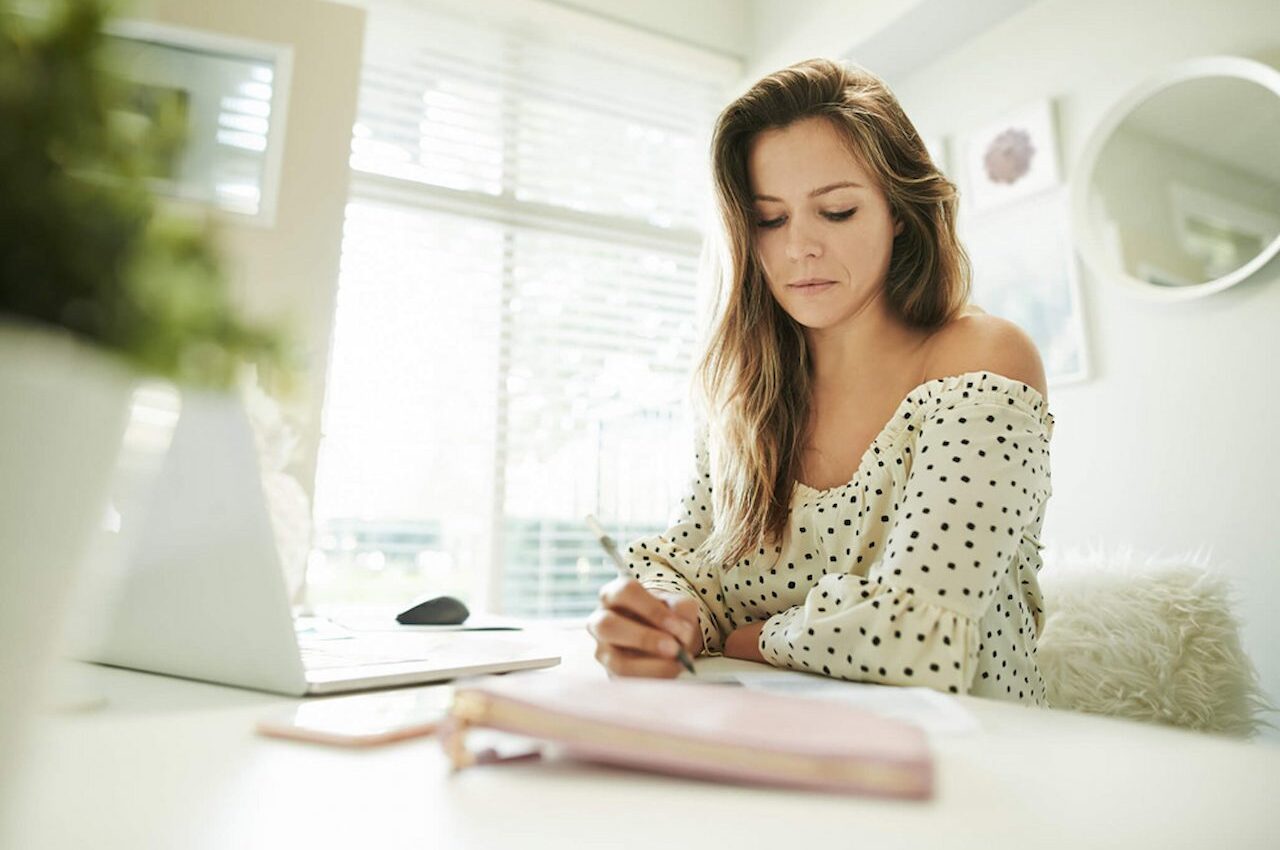 Mulher com expressão concentrada, anotando gastos mensais no papel e com computador aberto a sua frente.