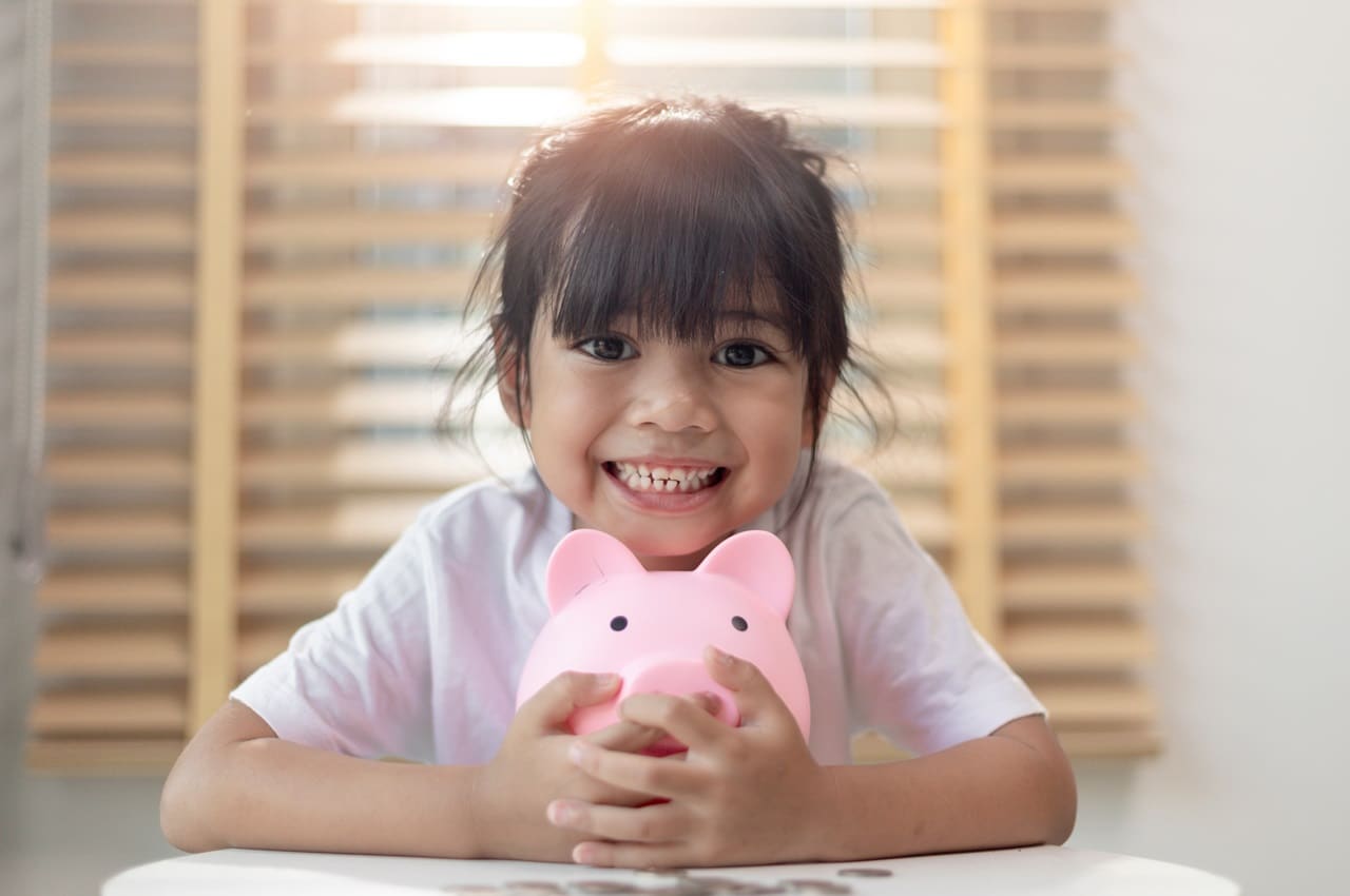 Criança sorridente de cabelos castanhos presos e franja, abraçando com as mãos um cofrinho em formato de porco e com moedas ao redor.