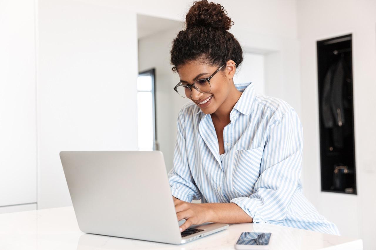 Mulher sorridente com os cabelos presos em coque, vestindo blusa de botões listrada e digitando no computador que está sobre uma bancada branca.