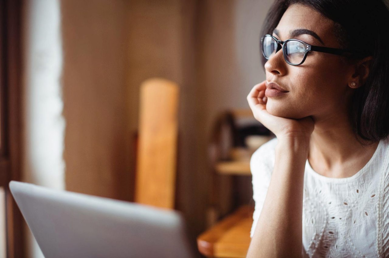 Mulher com expressão concentrada digitando em um computador. Ela usa óculos de grau e blusa básica branca.