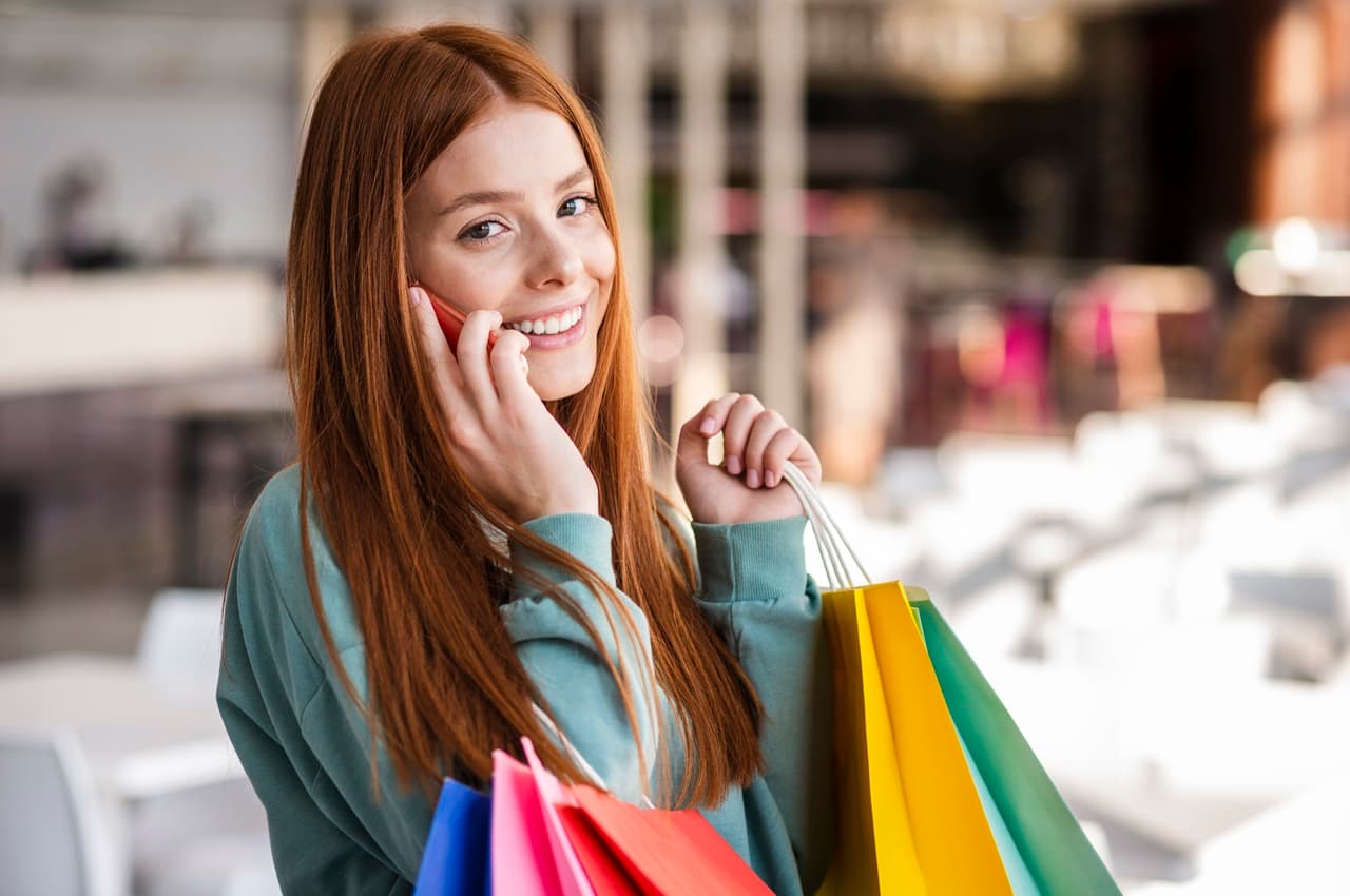 Mulher sorridente de longos cabelos ruivos, segurando sacolas de compras coloridas no dia do cliente e falando ao celular.