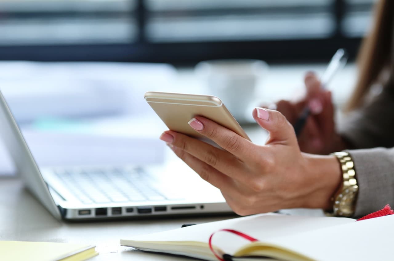 Mulher no ambiente de trabalho segurando celular e conferindo no computador aumento salarial. Ela veste terno cinza, relógio de ouro e seu rosto não aparece na imagem.