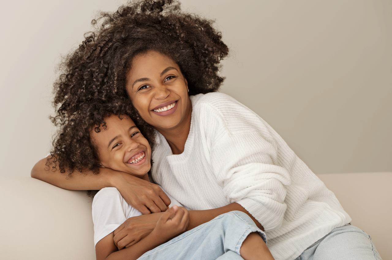 Família sorridente, composta por um menino pequeno e uma mulher, ambos vestindo calça jeans clara e blusa de lã branca.