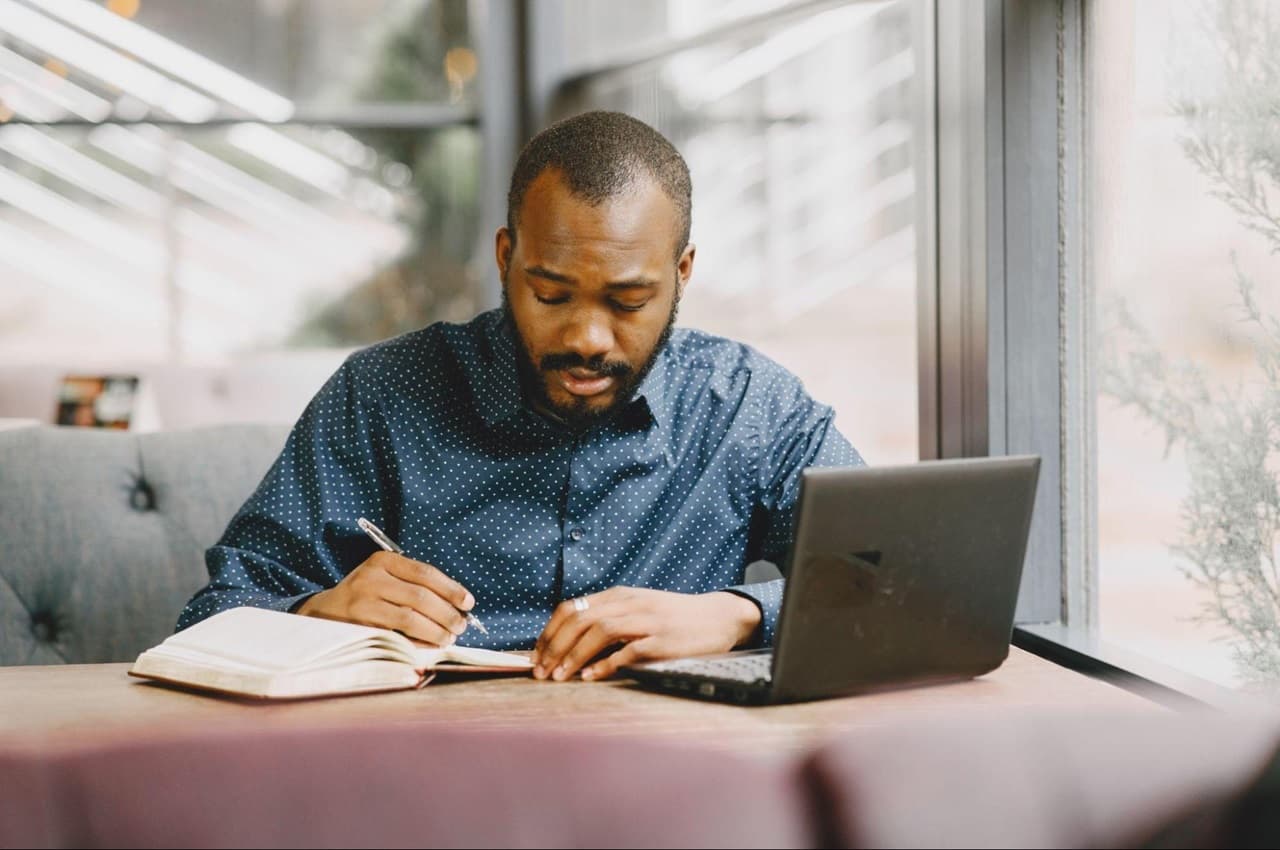 Homem com expressão concentrada e blusa social azul escura, anotando dicas financeiras para sair do vermelho em um caderno, enquanto utiliza um computador a sua frente.