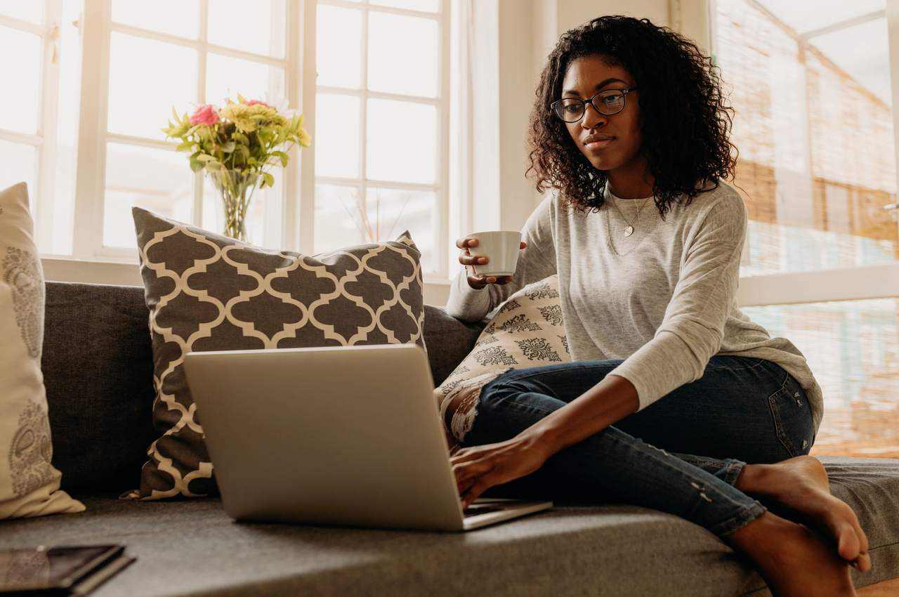 Mulher negra com cabelos cacheados soltos, sentada no sofá de casa e utilizando computador para dicionário financeiro.